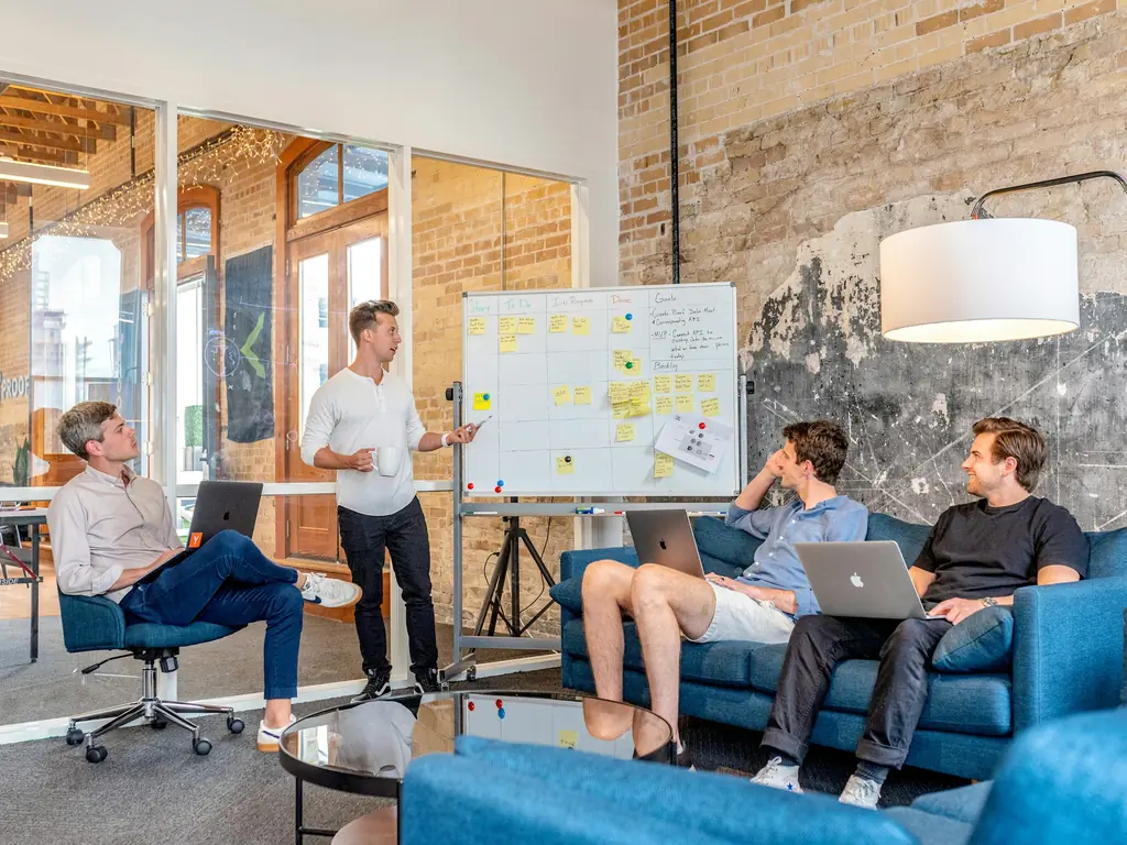 A team on sofas with laptops reviewing sticky notes on a whiteboard